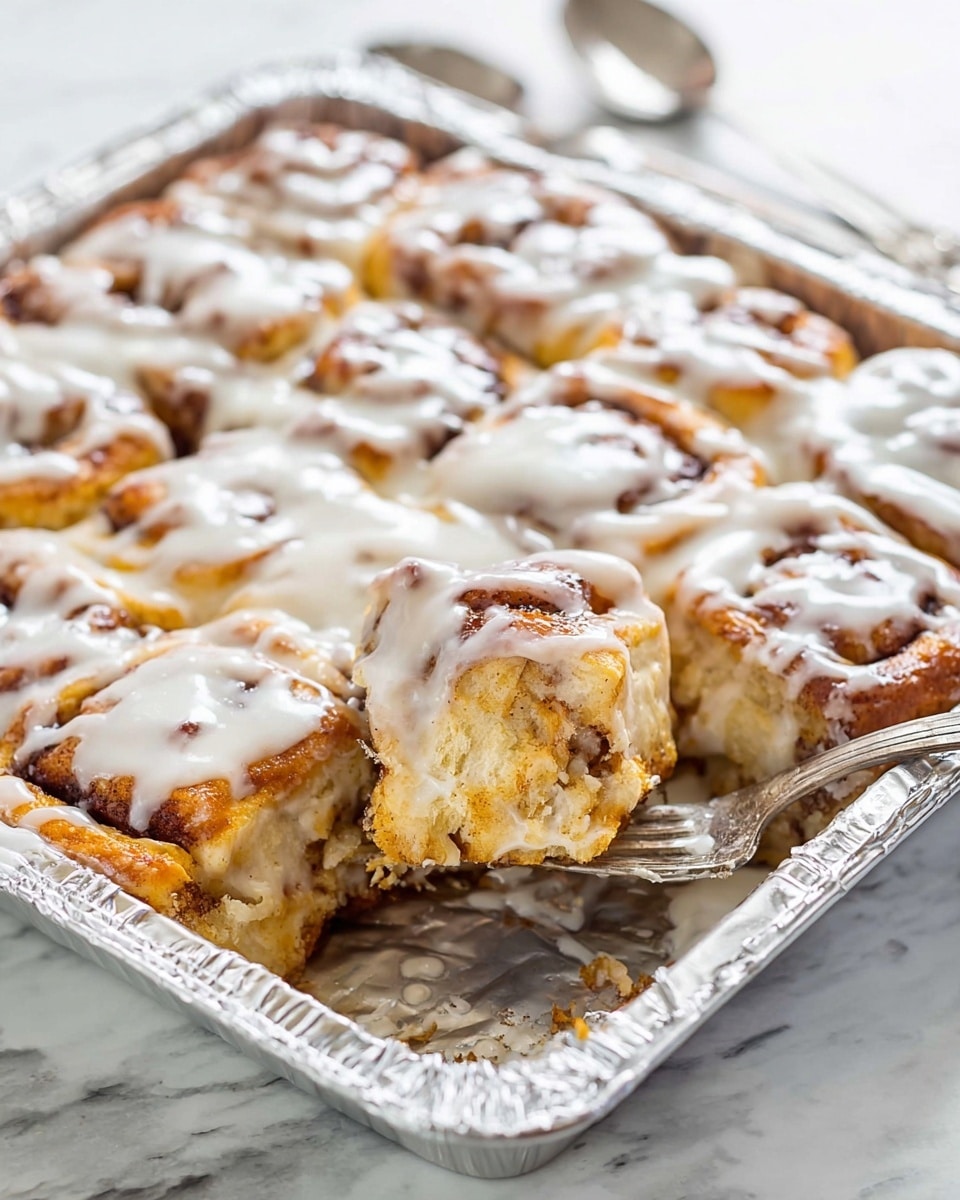 The image shows a square baking pan lined with shiny silver foil, filled with golden-brown cinnamon roll pieces stacked in layers, each topped generously with thick white icing that drips slightly over the edges. A corner piece has been cut out and is being held by a silver fork resting inside the pan, showing the soft, fluffy inside of the cinnamon rolls with visible cinnamon swirls. The background is a white marbled surface with a silver spoon blurred in the back. photo taken with an iphone --ar 4:5 --v 7