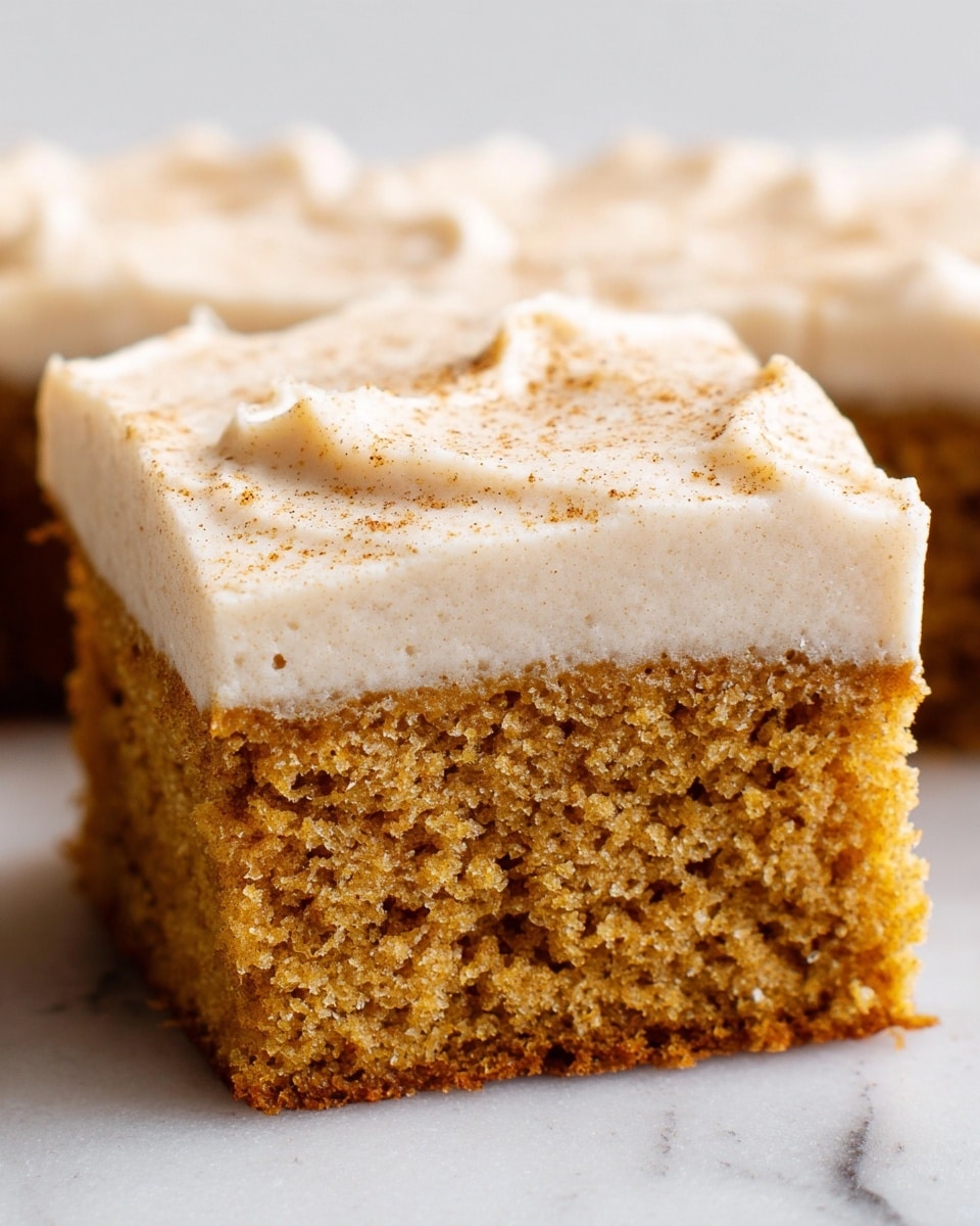The image shows a close-up of a single square piece of cake with two layers; the bottom layer is a moist, crumbly cake in a warm brown color with a slightly coarse texture, and the top layer is a thick, smooth, light beige frosting with a few tiny dark specks on the surface, spread evenly with soft peaks and swirls. The cake piece sits on a flat white marbled surface, and in the background, blurred similar pieces of cake can be seen. photo taken with an iphone --ar 4:5 --v 7