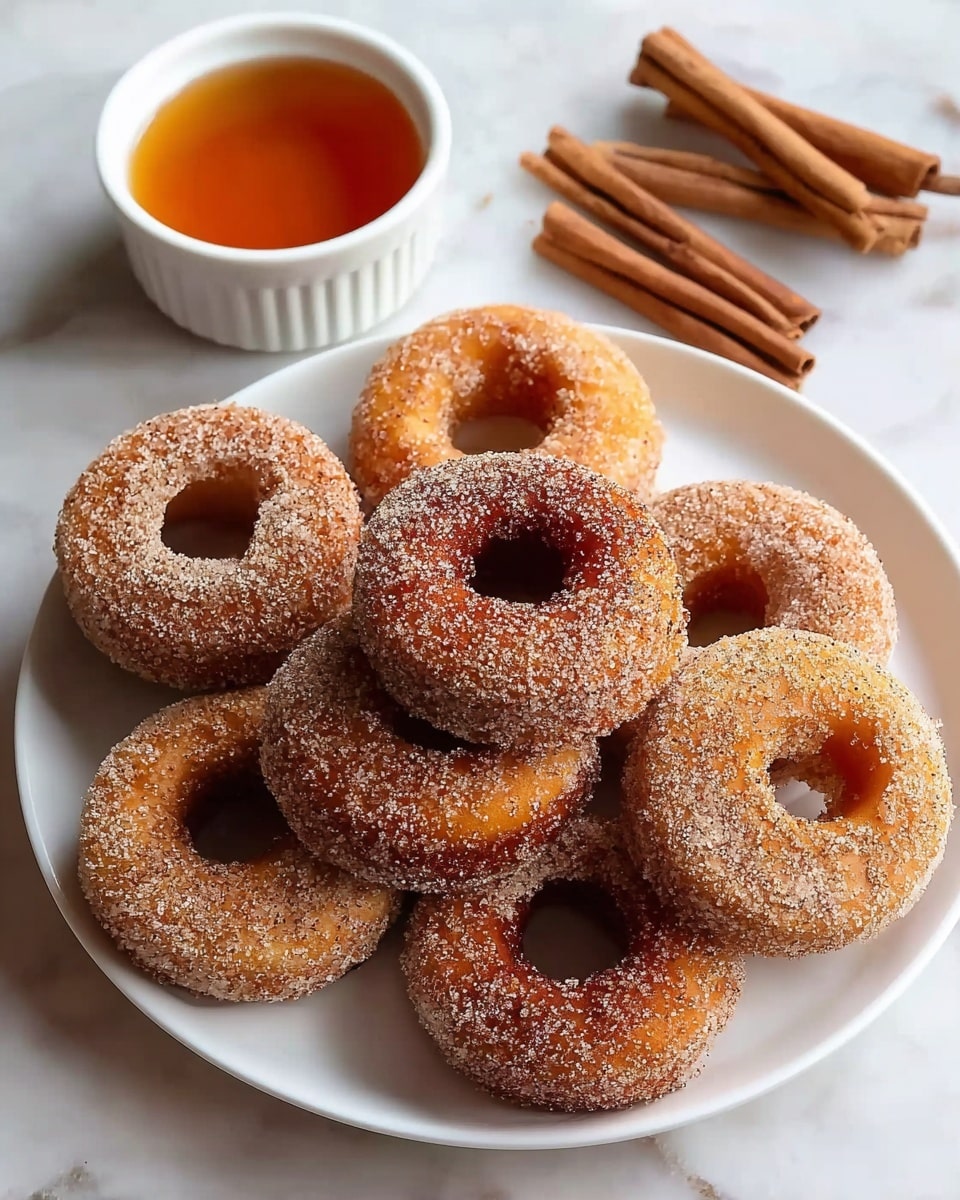 A white plate holds seven cinnamon sugar-coated donuts, each donut showing a rough texture with granulated sugar grains sparkling against the golden-brown fried dough. The donuts are stacked closely, some tilted to reveal light brown interiors through their circular holes. To the upper left on a white marbled surface sits a white ramekin filled with amber-colored syrup, with cinnamon sticks arranged in small bundles nearby. The white marbled background adds a clean and soft contrast to the warm tones of the donuts and cinnamon sticks. photo taken with an iphone --ar 4:5 --v 7
