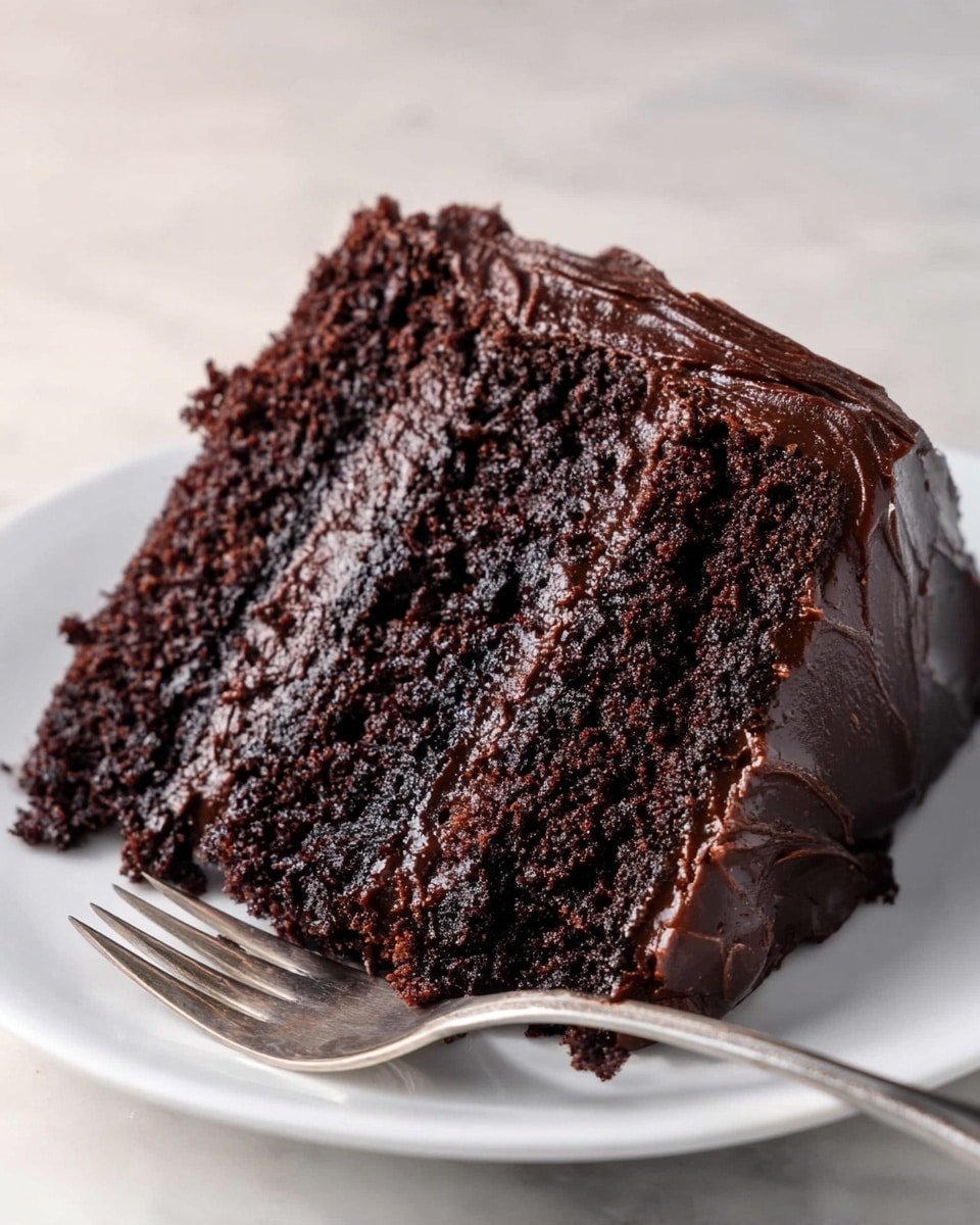 A close-up view of a three-layer chocolate cake slice placed on a white plate, showing dark, moist, and dense cake layers separated by thick, glossy dark chocolate frosting. The outside edges of the slice are fully coated in rich, smooth dark chocolate frosting with slight swirled texture. A silver fork rests near the front edge of the plate on a white marbled texture surface. photo taken with an iphone --ar 4:5 --v 7