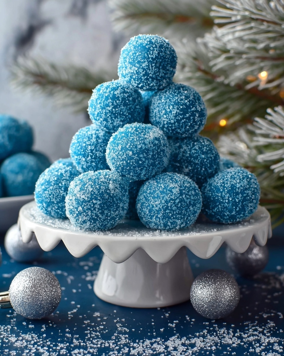 A stack of ten bright blue round sweets covered with coarse white sugar crystals, piled in a pyramid shape on a white, scalloped-edge cake stand; the sugar on the sweets sparkles under soft light, and the stand sits on a dark blue surface with scattered sugar crystals and silver glitter balls nearby, with some pine branches dusted with sugar in the background, all against a white marbled texture. photo taken with an iphone --ar 4:5 --v 7