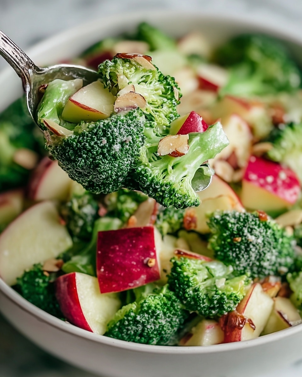 The image shows a close-up of a fresh salad with three main layers: bright green broccoli florets with a slightly creamy texture on the surface, chunks of red-skinned apples showing a mix of shiny red skin and pale flesh, and thin, light brown almond slices scattered throughout. The ingredients look fresh and mixed together closely, with small bits of dressing visible on both broccoli and apple pieces. The salad is in a white bowl placed on a white marbled surface. A spoon is lifting a portion, showing the detailed texture of each element. photo taken with an iphone --ar 4:5 --v 7