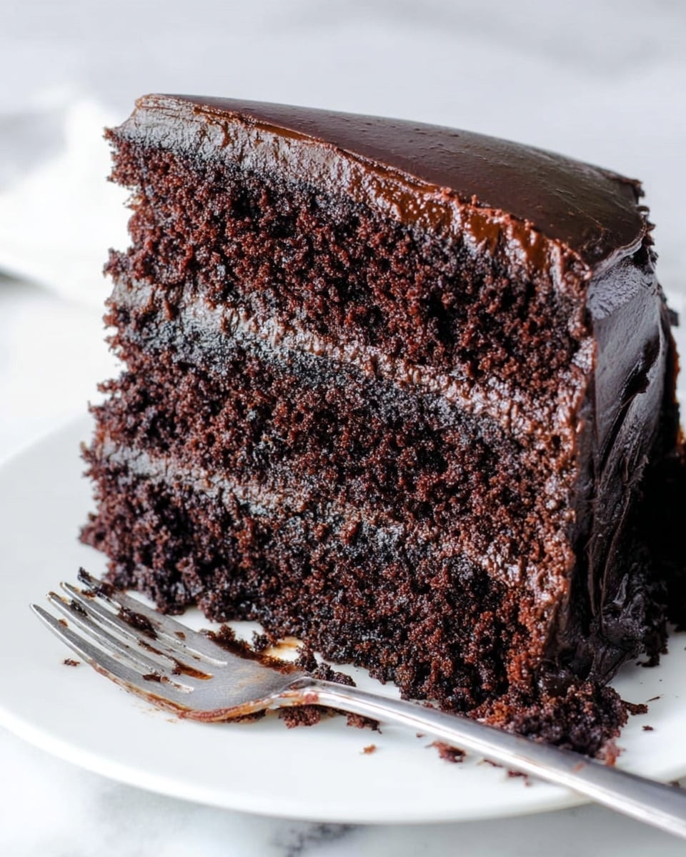 A close-up of a thick slice of rich, dark chocolate cake with three visible layers separated by glossy, dark chocolate frosting. The cake layers look soft and moist with a slightly crumbly texture. The smooth and shiny frosting covers the sides and top edge of the slice. The piece rests on a white plate with a slightly messy silver fork lying in front of the slice. The background is a white marbled surface. Photo taken with an iphone --ar 4:5 --v 7