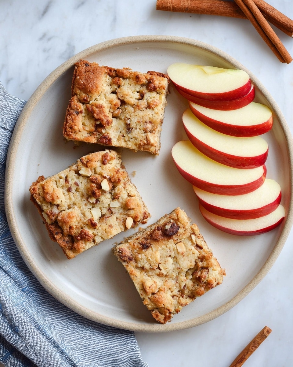 The image shows a white plate on a white marbled surface with three square pieces of crumbly apple and nut cake arranged on the left side. The cake has a light golden-brown crust with visible chopped nuts and small apple chunks embedded throughout. On the right side of the plate, there are five thin slices of red-skinned apple, fanned out neatly. The plate is next to some cinnamon sticks and a blue-striped cloth partially visible in the corner. photo taken with an iphone --ar 4:5 --v 7